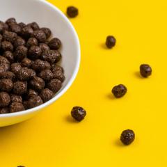 White bowl with chocolate corn cereal balls and milk on yellow background, top-down view.