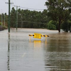 A road block with a sign that reads “WATER OVER ROAD” blocks a flooded road. 