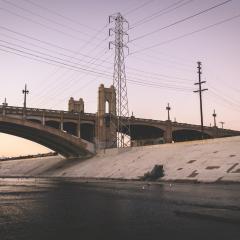 Los Angeles River with concrete sides, a bridge and energy pylons surrounding it against a dusk sky.