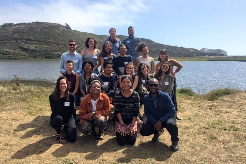 A group photo of Switzer Fellows on the beach with water, hills and a blue sky in the background.