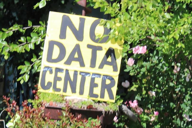 A yellow sign with black text reading “NO DATA CENTER” is nestled amid a planter of flowers in a garden in Kansas.  
