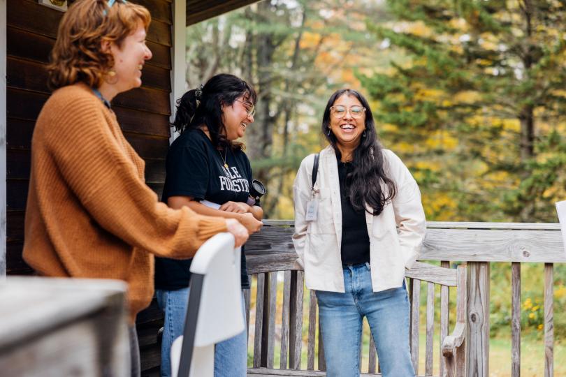 Three people are standing on an outdoor deck, laughing. There are fall leaves in the background. 