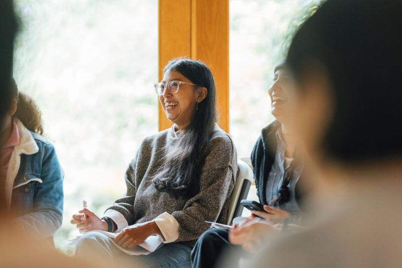 A Switzer Fellow is smiling and taking notes during the 2024 Switzer Fall Retreat. The background is a wall of windows with sunlight coming in. In the foreground are blurry heads, framing the fellow.