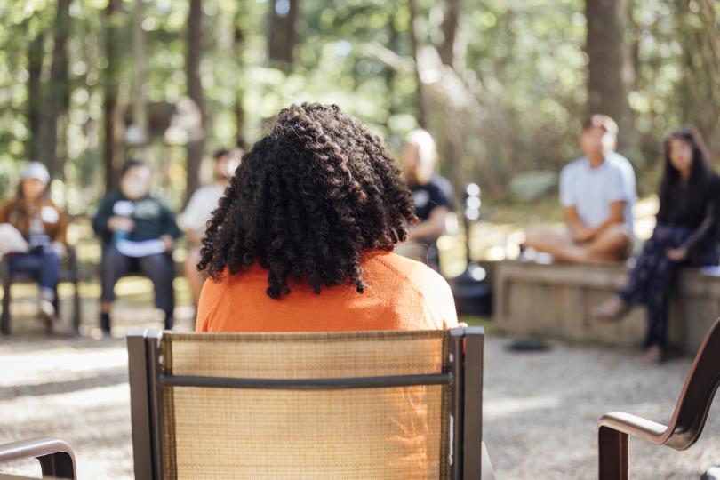 The back of a person's head, with curly, black hair and an orange shirt, sitting in a outdoor circle of people sitting in chairs, with trees and sunlight in the background.
