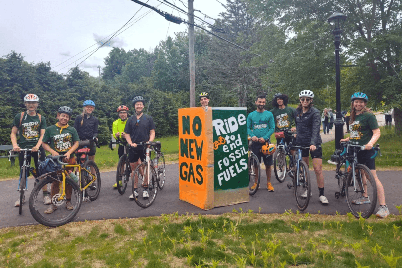 A group of cyclists stand with their bikes, smiling. A green and orange sign in the middle reads 'no new gas' and 'ride to end fossil fuels'.