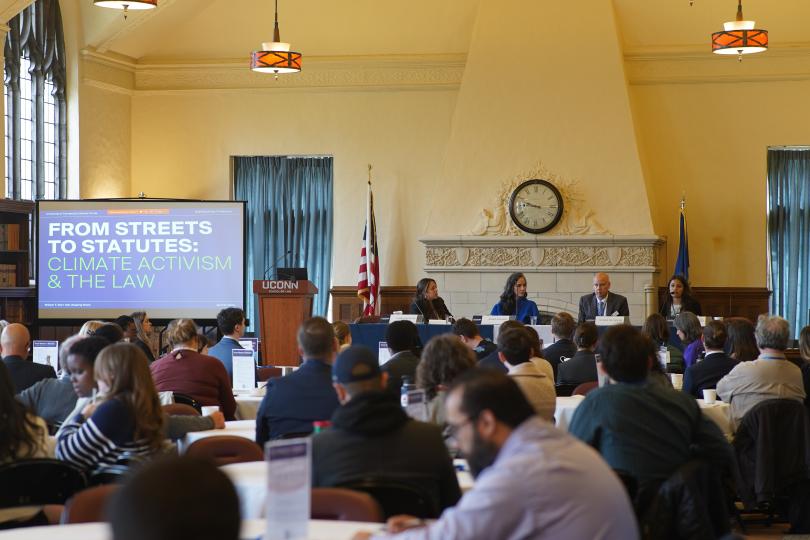 Four speakers sit at a table at the front of a room with a powerpoint and a full audience of people