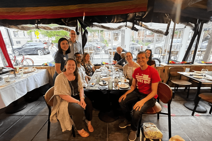 Switzer Fellows sit and stand around a restaurant table smiling. 