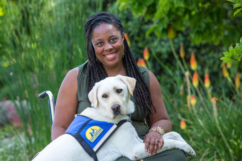 India Harville sits in a wheelchair with her white Labrador service dog, Nico, on her lap. They are outside with green plants in the background.