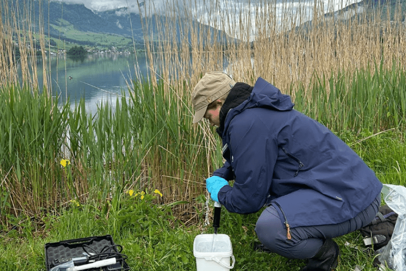Kristy crouches in the grass next to a lake in the mountains. She is using a eDNA monitoring kit, pumping water out of a white bucket through a black pipe, with other sampling materials in bags and containers on the grass nearby.