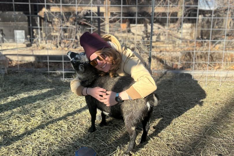 Maya Higgins crouches, wearing a maroon winter hat, hugging a small grey and black goat on her home farm. There are farm structures and plants in the background.