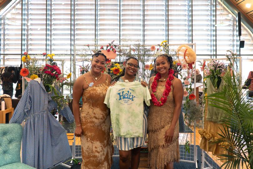 Mica and Maya stand with another person in front of a display of flowers and clothing, holding a tie dye t-shirt that reads "NO WAR, NO ICE, HELIX FOR LIFE, New Haven CT, April 17 2026" in blue text.