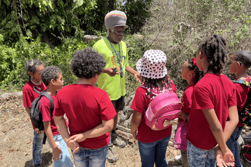 Students in red t-shirts stand listening to a man in a yellow shirt and knitted hat speaking outside at the St. George Village Botanical Garden as he discusses the slave grave site located within the gardens.