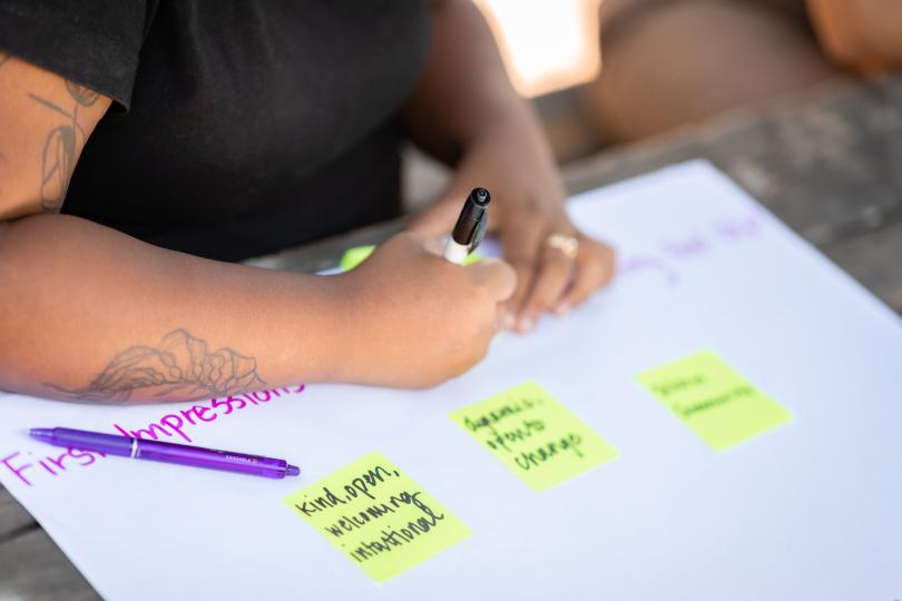 A close up of hands writing on post it notes against a white paper background with "First Impressions" written on it, and already written post it notes reading "kind, welcoming, open, intentional"