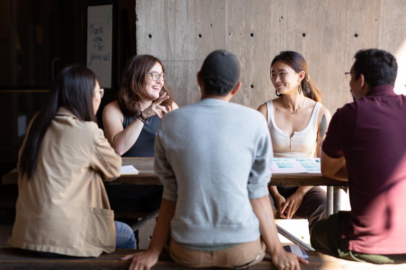 A group of five fellows sitting together at a picnic table smiling at one another.