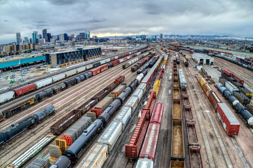 Aerial view of a trainyard with many train tracks, some full of different kinds of trains and others empty. The Denver, CO skyline is in the background under a grey cloudy sky.