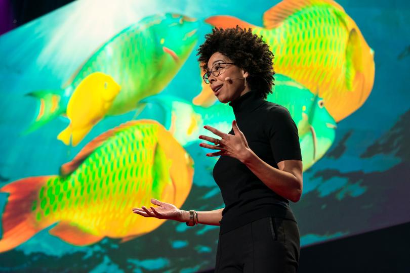 Ayana Elizabeth Johnson is seen on stage from below, with a screen showing colorful fish on it as the background. She is wearing a microphone, a black short-sleeved turtleneck and holding both hands out as she speaks.