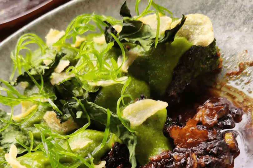 A close up of a meal created from all parts of a broccoli plant. It is bright green with a brown sauce on a white plate.