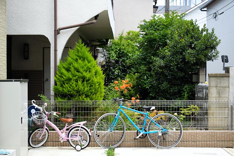 A pink child's bike and a blue adult's bike rest on a fence near concrete buildings and manicured green trees. 