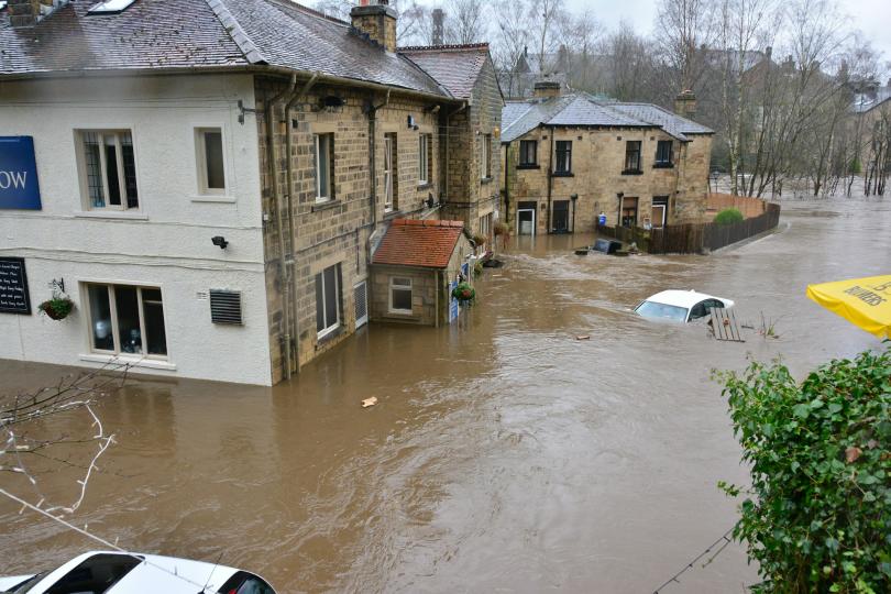Brown water floods a street in Ireland. A white car is mostly submerged in the water, and the water is halfway up the doors to the buildings on the street. The buildings are made of stone bricks with tile roofs. 
