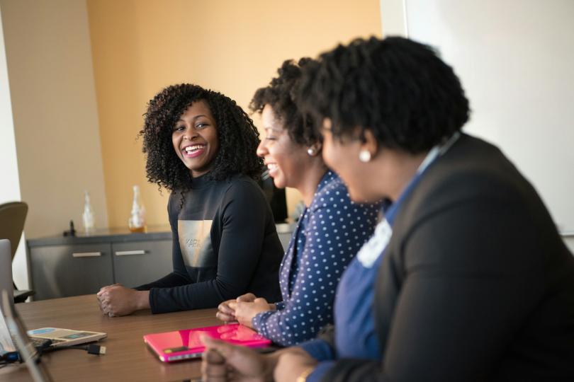 Three Black women sit at a meeting table next to each other smiling and laughing. 