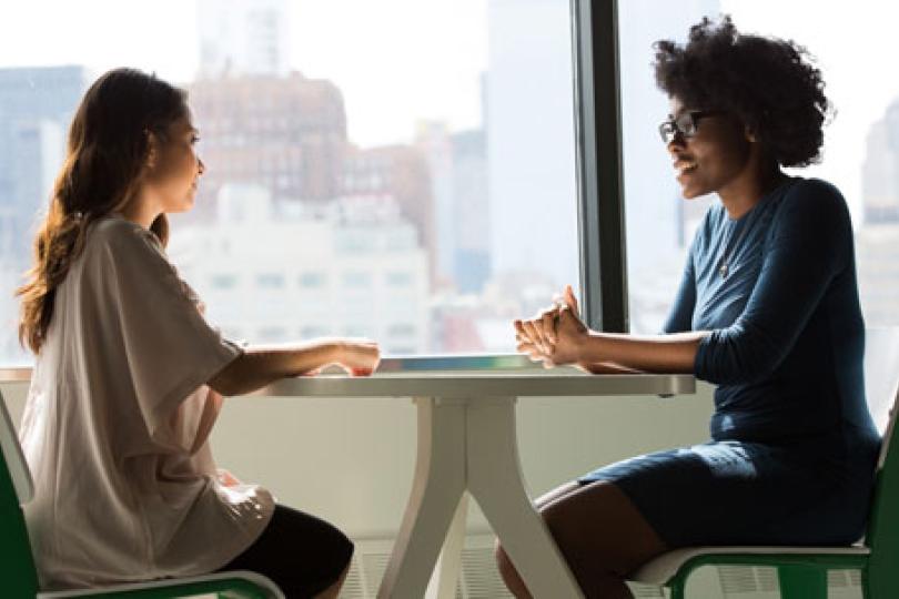 Two women sitting at a tall table next to windows with city buildings in the distance.