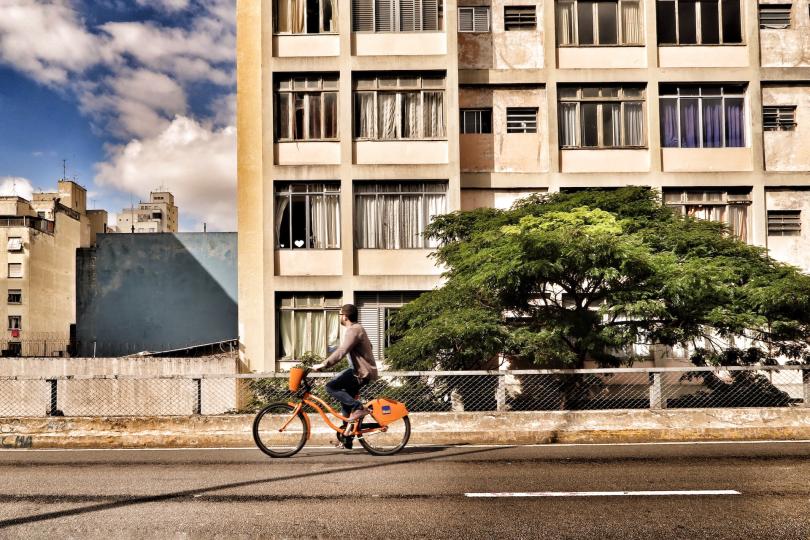 A person is biking on a road with a tree and a concrete apartment building in the background.
