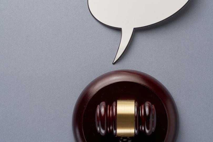 A brown legal gavel sits in a holder on a grey background. An empty white speech bubble sits above it.