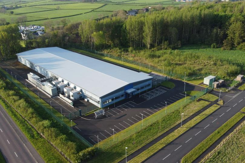 An aerial view of a Data Centre in Coleraine, Ireland. It’s a large white building with a small parking lot. There are small roads, green grass, woods, and a farm in the surrounding area. 