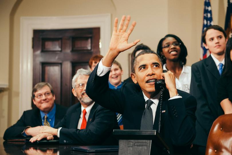 President Obama sits at a desk in the White House, holding a black phone and waving while looking up and to the side. Senators sit with him and middle school students stand behind them.