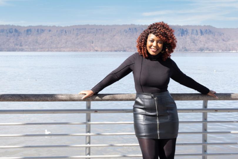 Janelle is smiling and standing in front of a railing with water and hills in the background