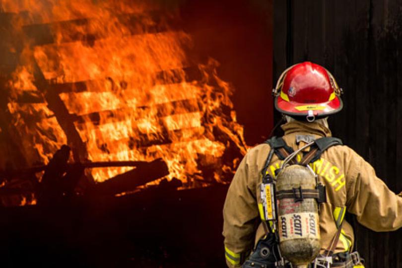 A firefighter stands facing away from the camera, toward a structure burning with bright orange flames