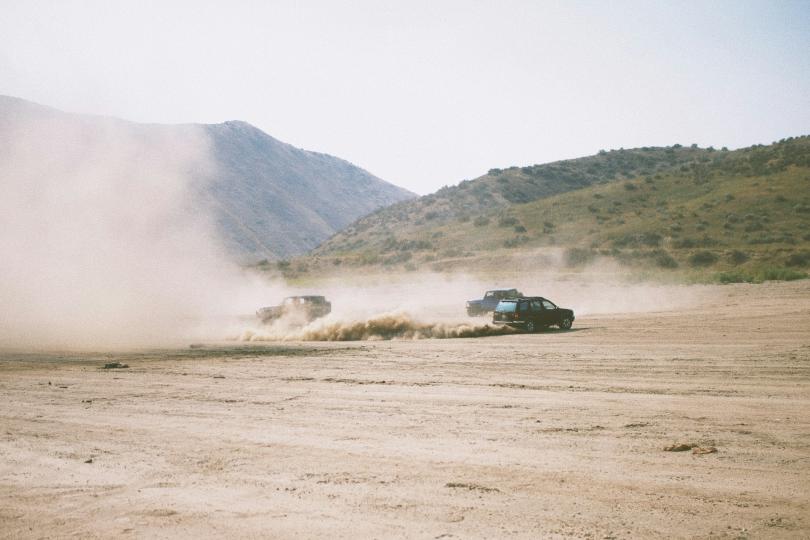 Two jeeps and a car drive on a dusty flat landscape with green hills in the background, kicking up a large dust cloud.