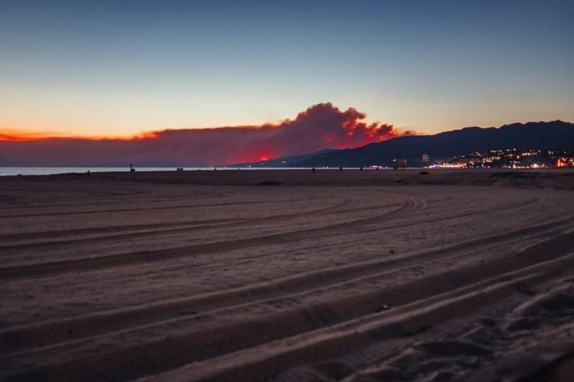 A ground level photo shows beach sand in the foreground, with a coastline and hills with city lights at dusk in the background, with a huge cloud of grey wildfire smoke backlit with the orange of the sunset.