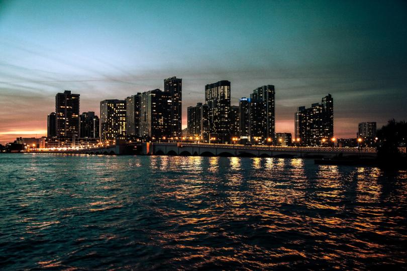 Sunset behind tall buildings along the Miami, FL coast, seen across water.