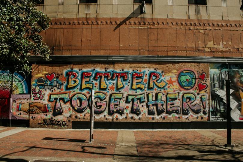 A mural is spray painted onto a building that is boarded up with plywood. The mural reads BETTER TOGETHER! with hands holding each other, a globe, red hearts, and a city skyline.  