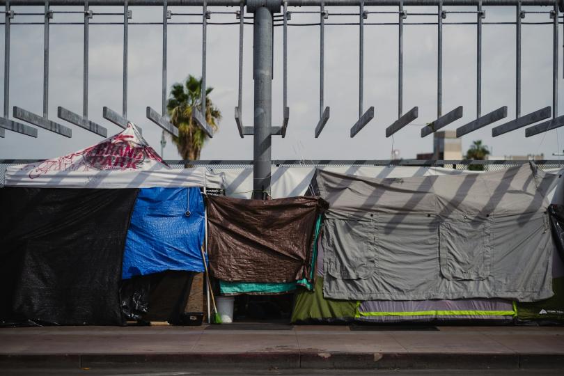 A homeless encampment is made up of tarps and tents set up neatly under a metal structure with hanging bars. Open sky with clouds, a couple buildings and a palm tree are in the background. 