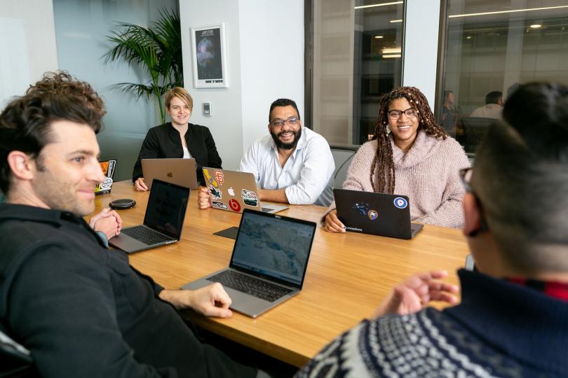 A group of people sit around a table with laptops open at a meeting, smiling and laughing with each other.