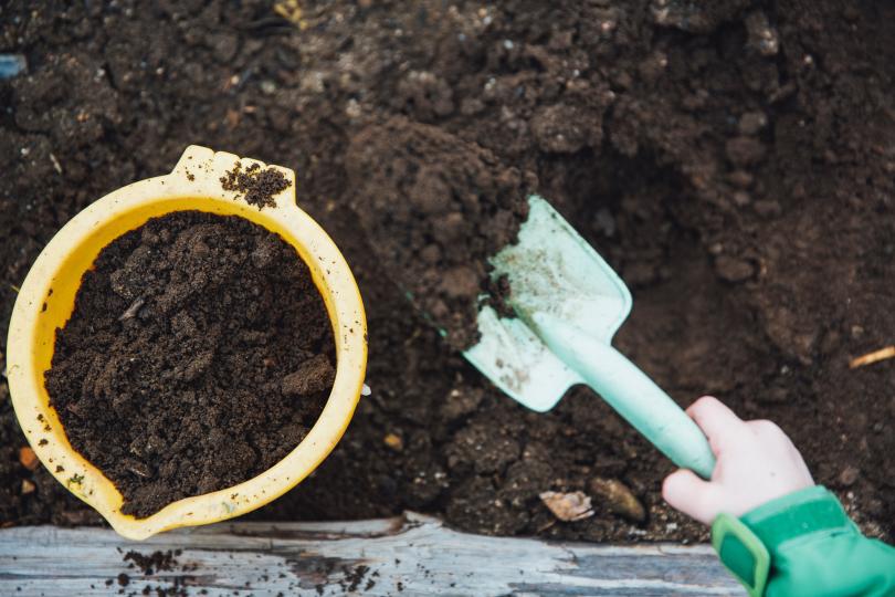 A child's hand scoops brown soil into a yellow pot with a gardening trowel. 