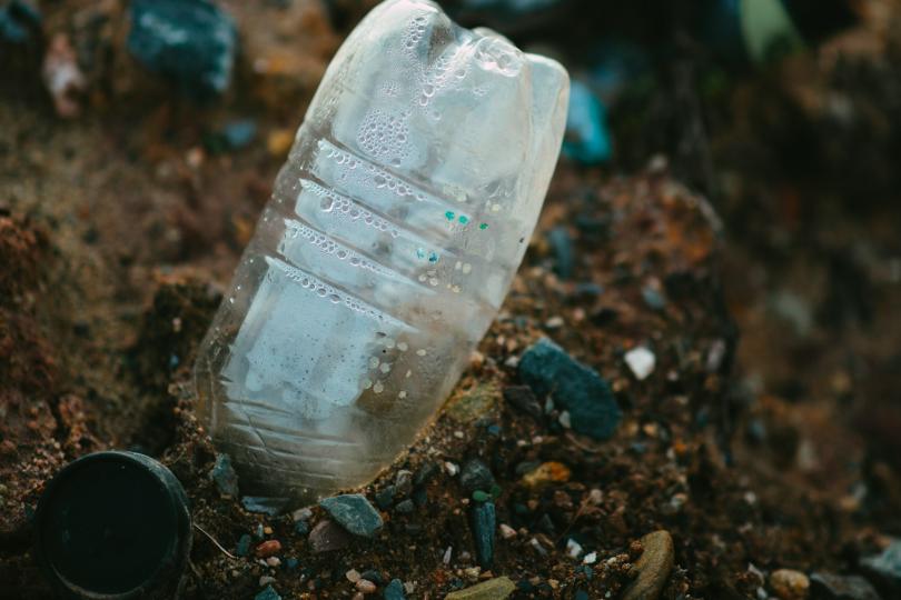 A plastic water bottle pokes out of brown soil dotted with rocks, with other plastic trash out of focus in the background. 