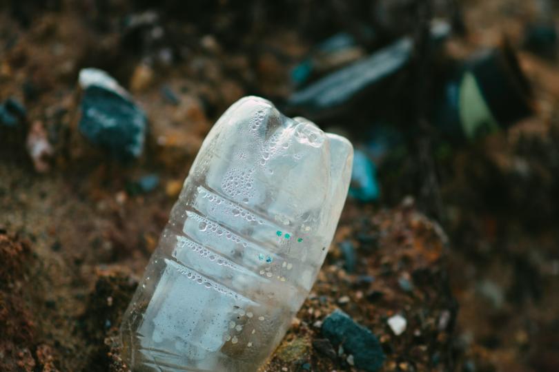 A plastic water bottle pokes out of brown soil dotted with rocks, with other plastic trash out of focus in the background. 