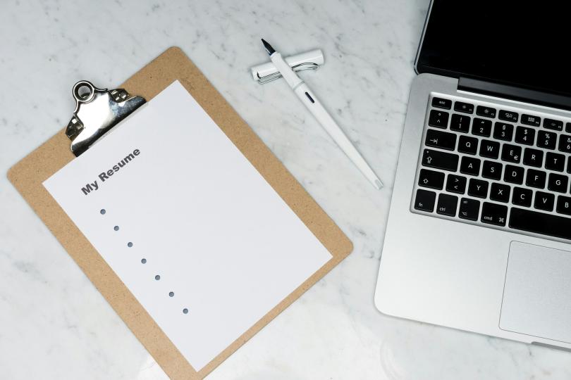 A clipboard with a resume template, a pen and a mac laptop sit on a white marble table.