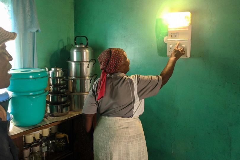 An African woman in a headscarf and apron faces away, turning on an electric wall lamp mounted on a green wall in a kitchen. Pots and pans are on a counter behind her and the profile of another person's face is watching and speaking, cut off on the left.