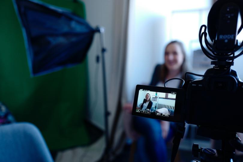 A woman is being filmed by a video recording camera, seen from behind the camera.