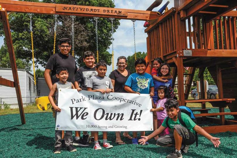 A group of children and adults stand in front of a playground holding a white banner that says "We Own It!"