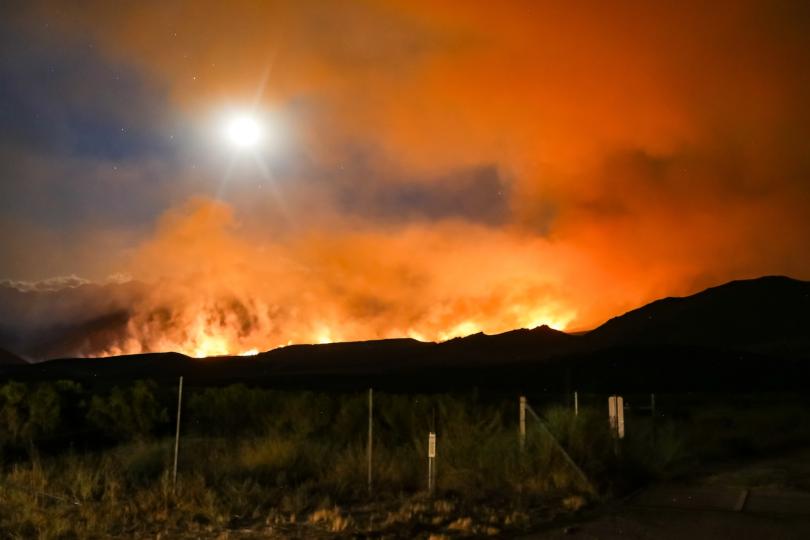 Fires burn over the crest of a dark hill with orange smoke rising.