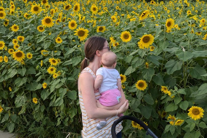 Cora stands holding her daughter, looking away from the camera into field of sunflowers.