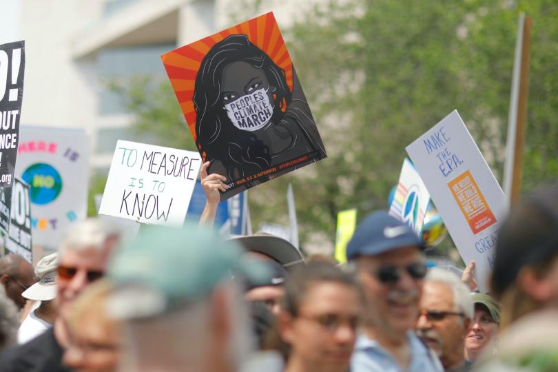 At a climate march, a hand holds up a sign of showing a Black woman against an orange background, wearing a white mask that reads PEOPLES CLIMATE MARCH.