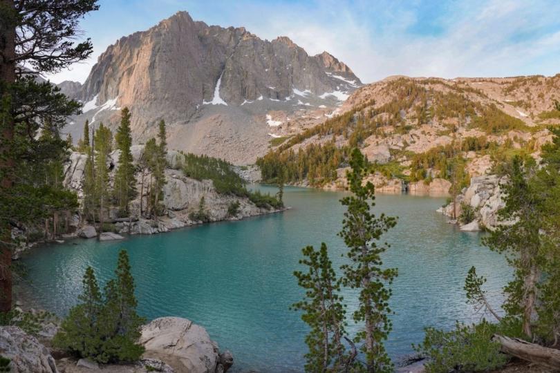 Pine trees and rocks surround a turquoise blue lake with a mountain and blue sky in the background. 