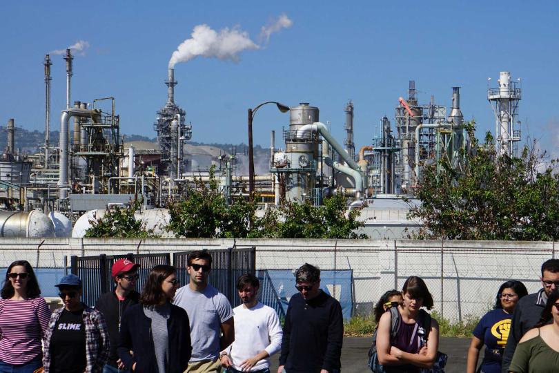 UCLA Law students and faculty standing and walking in front of an energy plant full of machinery and smokestacks, with a blue sky in the background during a toxic tour of Wilmington, CA, a predominantly Latinx neighborhood surrounded by industry and oil.