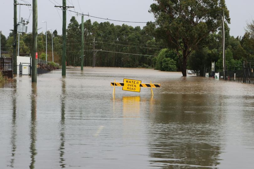 A road block with a sign that reads “WATER OVER ROAD” blocks a flooded road. 